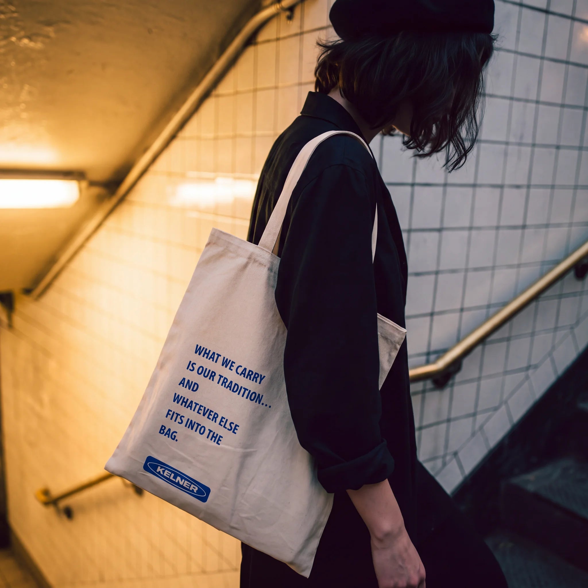 Person carrying the canvas KELNER tote with its What We Carry statement on a tiled subway stairway